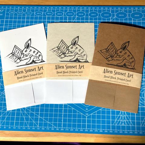 a set of three cards laid out on a blue cutting mat. a brown Kraft card, a plain white card, and a cream parchment pattered card. each card has a yellow parchment strip of paper wrapped around it that says "Alien Sunset Art" Hand Block Printed Card.

the block print on the cards is of  a bunny reading a book