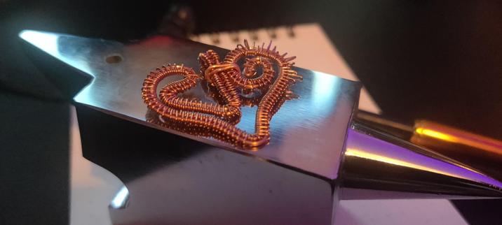 a wire-wrapped pendant in the shape of the Crest of Love from Digimon adventure. It is made of copper wire and lies on a steel anvil. A mallet is laying in the background