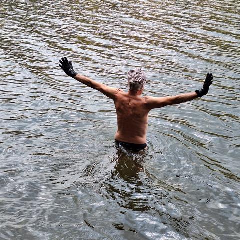 Ein Mann mit schwarzer Badehose und gestreifter Mütze steht bis zur Lende im Wasser. An den Händen trägt er Neoprenhandschuhe. Die Arme sind seitlich leicht nach oben ausgestreckt. Das Wasser bildet diagonale Wellen, um den Mann einen kreisförmigen Strudel.
Es ist blassblau, hell- und dunkelgrau und zeigt sich im Wechselspiel mit Hell und Dunkel.