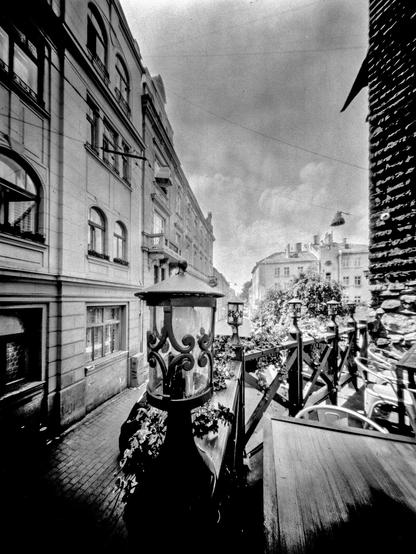The image depicts a quiet street scene in Lviv, captured with a pinhole camera. In the foreground, there is an ornate metal lantern surrounded by plants and a wooden railing, possibly part of a café terrace. The narrow street stretches into the distance between old European-style buildings, their facades showing classic architectural details. The image has a soft, slightly dreamlike quality, characteristic of pinhole photography, with gentle light diffusion and a wide depth of field that emphasizes both the foreground textures and the receding perspective of the street