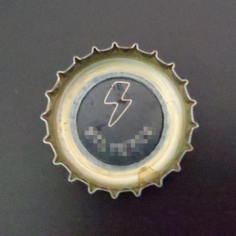 Zu sehen ist die Innenseite eines Kronkorkens auf schwarzem Hintergrund.
Im Kronkorken ist eine Zeichnung eines Blitzes und ein verpixelte Schrift zu sehen. Die Schrift ist nicht gerade sondern verläuft im Halbkreis unterhalb um den Blitz