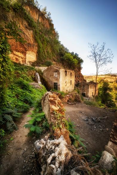 Verfallene Häuser am Fuss einer steilen, rötlich-braunen Felswand, umgeben von dichtem Grün aus Farnen und Sträuchern. Ein schmaler Erdpfad führt an einer halb eingestürzten Mauer vorbei. Durch eine Öffnung in der Felswand fliesst ein kleiner Wasserfall neben einem der Gebäude herab. Im Hintergrund weitet sich die Landschaft unter einem klaren blauen Himmel, ein einzelner kahler Baum steht am Rand des Geländes.
