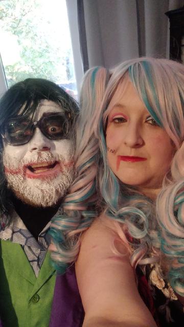 Alt text:
A couple dressed as the Joker and Harley Quinn pose together. The Joker has white face paint, dark eyes, and red lipstick smeared into a grin, while Harley Quinn wears a pastel pink and blue wig with matching makeup and a playful expression. The lighting is warm and playful.