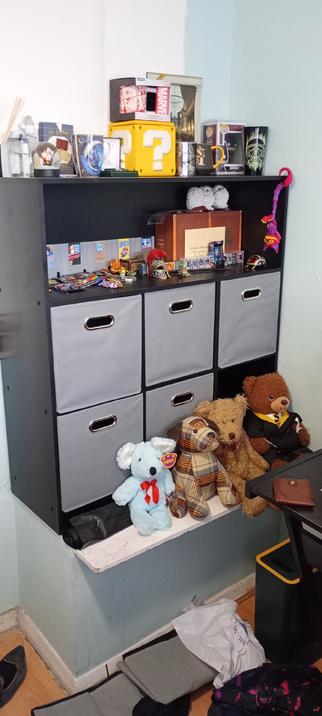 Alt text:
A black shelving unit filled with collectibles and memorabilia. The top shelves feature items like a yellow Mario question block, Marvel and Batman mugs, Harry Potter merchandise, and small figurines. The lower shelves contain grey fabric drawers, with teddy bears and plush toys arranged neatly below.
