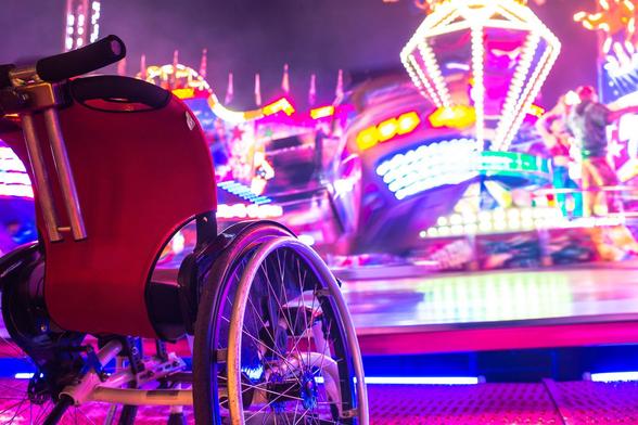 Ein leerer Rollstuhl steht auf einem Jahrmarkt, vor einem sich drehenden Karussell. Das Fahrgeschäft hat Bewegungsunschärfe. Alles ist sehr bunt auf dem Foto.