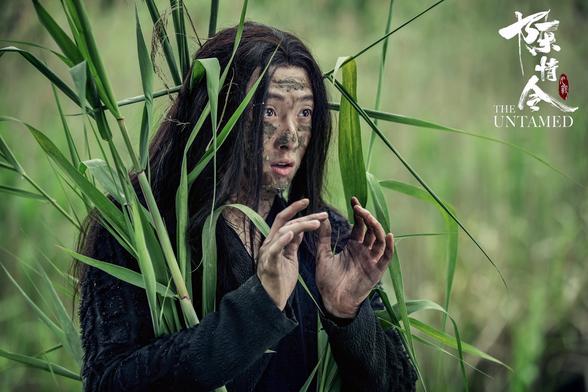 Wen Ning, dressed in black with mud smudged over the exposed skin of his face, neck, and hands, and with long green plant stalks strapped around his body for bleeding in with the field of tall green behind him. His hands are raised to chest level, caught in the moment of explaining to someone offscreen what he's been up to.