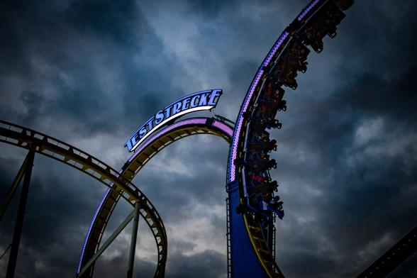 Eine Achterbahn im Sturm. Auf dem Looping steht das Wort ,Teststrecke‘ im blauen Neonbuchstaben, die sich gegen den dramatischem Wolkenhimmel absetzen.