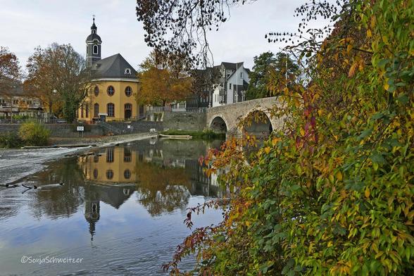 Die gelbe Hospitalkirche spiegelt sich in der Lahn, rechts von ihr sieht man ein Stück der alten Lahnbrücke, neben der Lahnbrücke im Bild, sind herbstlich gefärbte Blätter zu sehen.