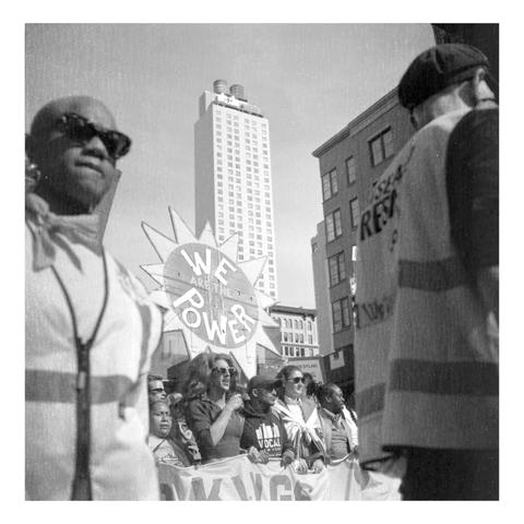 A Line of protesters, framed by 2 individuals in the foreground, with a visible sign shaped as a sun, with the words “We Are The Power”.