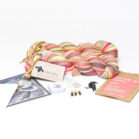Picture shows two skeins of yarn, one behind the other, in shades of black, yellow, red and coral. In front of the yarn, from left to right, is a card, a tin of lip balm, cat stitch markers, a crow sticker and a packet of tea.