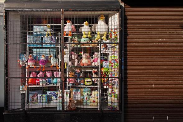 Closed gift shop with brown metal shutters and a grill over a window containing faded toy boxes and a range of cuddy toys mostly of the seagull stealing a chip or hotdog variety, low sun and shadows