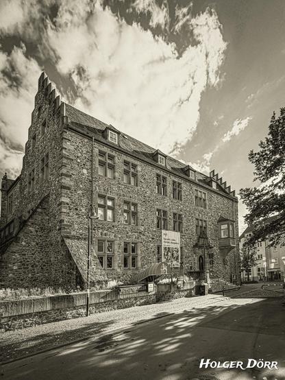 Ein historisches Gebäude aus hellem Bruchstein erhebt sich mächtig unter einem teils wolkigen Himmel. Die Sonne taucht die Fassade des Alten Schlosses in Gießen in warmes Licht und lässt die Struktur des Mauerwerks lebendig wirken. Die steilen Treppen, der spitze Treppengiebel und die markanten Fenster verleihen dem Bau Würde und Zeitlosigkeit. Die Aufnahme in nostalgischem Schwarzweiß lässt die Szene wirken, als stamme sie aus einer anderen Epoche – ruhig, eindrucksvoll und voller Geschichte.