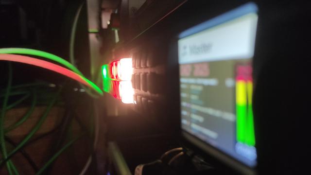 Foto eines im Rack verbauten Atem 2 M/E Constellation Videomischers, aus einem interessanten Winkel. Im Vordergrund ist unscharf das Display des Mischers zu sehen, weiter hinten im Bild leuchten verschiedene Tasten des Mischers Warmweiß, Rot und Grün. Die Tasten beleuchten rote und grüne Kabel, die sich am Mischer vorbei schlängeln. im Hintergrund sind im Dunkeln noch weitere Kabel vor einer hölzernen Stufe zu sehen.