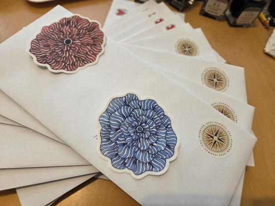 Fanned out stack of envelopes with international postage stamps. My floral art stickers are sitting on the addresses, for privacy of the recipient and myself.  There are little ink bottles next to the envelopes.