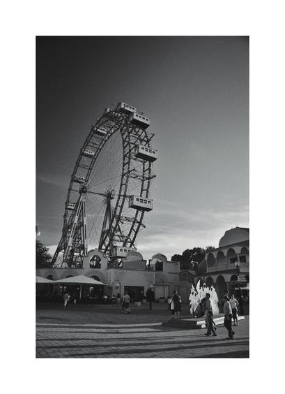 A low-angle, black and white photograph of a large Ferris wheel, the Wiener Riesenrad in Vienna, Austria, dominates the frame under a bright sky. The wheel is seen from the ground level. In the foreground, people are walking on a paved area near some buildings and a fountain is shooting several jets of water into the air. The sun is shining, casting long shadows.