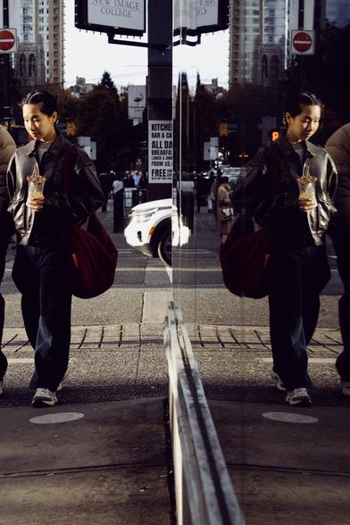 A woman, her face aglow, quietly looks down at the pavement. Her reflection does much the same.