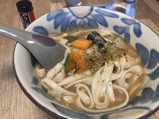 A bowl of noodle soup featuring wide, flat noodles, assorted vegetables like carrots and greens, and pieces of meat. A gray spoon rests in the bowl, and a small container of seasoning is visible beside it. The bowl has a decorative blue pattern,