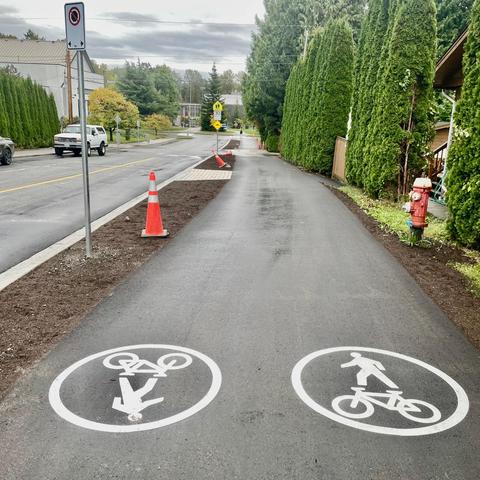A paved multi-use path on the south side of 19th street. In the foreground on the path are walking and biking symbols. In the background is Sutherland School.