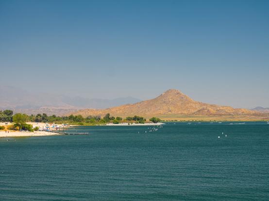 A pointy arid hill gradually rises above a vast artificial lake, where there are boats. More mountains are in the distance, in the haze.