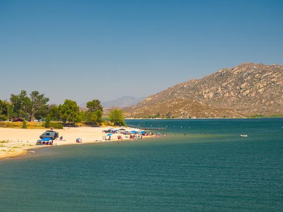 A beach and trees are by a vast artificial lake. Many people were playing on the beach and shallow parts of the lake. An arid hill rises above the lake in the distance.