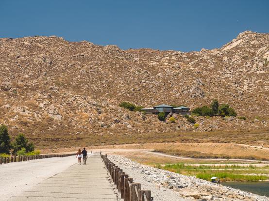 A couple were walking on the long dam top of Lake Perris. The dam top leads to a backdrop of an arid mountain. A modern building (Ya'i Heki' Regional Indian Museum) perches on the hill.