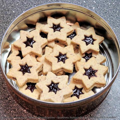 Auf der grauen Küchenplatte steht eine Blechdose mit hellen Haselnusssternen. In der Mitte ist ein sternförmiges Loch und die Marmelade guckt durch.