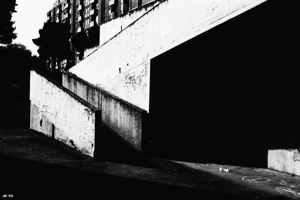 Lowkey-Schwarzweißfoto mit starkem Schwarzanteil. Es zeigt links den Aufgang zu einem großen Wohngebäude und rechts einen großen Bereich darunter, der komplett im Schatten liegt. Links oben im Hintergrund das mehrstöckige Gebäude selbst. Olympus 35LC auf Rollei IR400-Film.