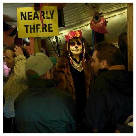 A sign reads Nearly There. The living dead looks through the lens between to lads chatting.