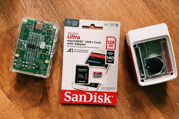 a photo showing three objects on a wooden table. From left to right: a RaspberryPi 3 in a clear case, a 128GB Sandisk microSD card still in its packaging and a RaspberryPi 5 in a white case