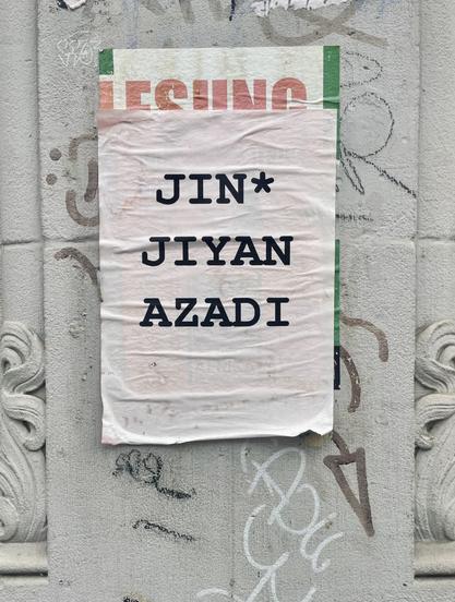 Ein verblichenes rosa Plakat auf einer grauen Hauswand, welche mit einigen Tags versehen ist. Auf dem Plakat in schwarz und in Caps mittig gesetzt „JIN* JIYAN AZADI“