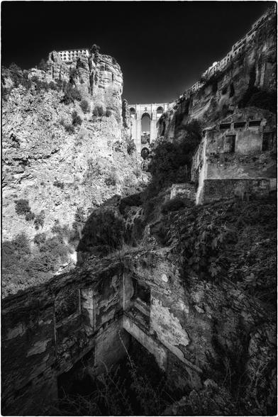 Ein tiefes, steiles Tal mit einer imposanten Brücke, die sich hoch über dem Abgrund spannt. Links ragt ein Felsen mit einem Gebäude darauf empor. Rechts und im Vordergrund führen alte, verwitterte Mauern und Ruinen den Hang hinunter. Die Szene wirkt dramatisch durch das Spiel von Licht und Schatten.