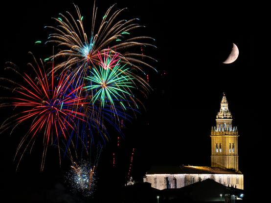 𝗣𝗶𝗰𝘁𝘂𝗿𝗲 𝗗𝗲𝘀𝗰𝗿𝗶𝗽𝘁𝗶𝗼𝗻 (𝗘𝗻𝗴): Nighttime photograph of fireworks over the sky of Nava del Rey. On the left, intensely colored fireworks explode: reds, greens, blues, and golds, forming different shapes and luminous trails. On the right side of the image, the illuminated tower of the Church of Santos Juanes stands out, its architectural details highlighted in warm tones. Above it, in the dark sky, the crescent moon is visible. 

𝗗𝗲𝘀𝗰𝗿𝗶𝗽𝗰𝗶𝗼́𝗻 (𝗘𝘀𝗽): Fotografía nocturna de fuegos artificiales sobre el cielo de Nava del Rey. En el lado izquierdo, estallan fuegos de colores intensos: rojos, verdes, azules y dorados, formando diferentes formas y trazos luminosos. En la parte derecha de la imagen aparece la torre iluminada de de la iglesia de los Santos Juanes, destacando sus detalles arquitectónicos en tonos cálidos. Sobre ella, en el cielo oscuro, se ve la luna en cuarto creciente.