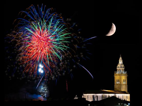 𝗣𝗶𝗰𝘁𝘂𝗿𝗲 𝗗𝗲𝘀𝗰𝗿𝗶𝗽𝘁𝗶𝗼𝗻 (𝗘𝗻𝗴): Nighttime photograph of fireworks over the sky of Nava del Rey. On the left, intensely colored fireworks explode: reds, greens, blues, and golds, forming different shapes and luminous trails. On the right side of the image, the illuminated tower of the Church of Santos Juanes stands out, its architectural details highlighted in warm tones. Above it, in the dark sky, the crescent moon is visible. 

𝗗𝗲𝘀𝗰𝗿𝗶𝗽𝗰𝗶𝗼́𝗻 (𝗘𝘀𝗽): Fotografía nocturna de fuegos artificiales sobre el cielo de Nava del Rey. En el lado izquierdo, estallan fuegos de colores intensos: rojos, verdes, azules y dorados, formando diferentes formas y trazos luminosos. En la parte derecha de la imagen aparece la torre iluminada de de la iglesia de los Santos Juanes, destacando sus detalles arquitectónicos en tonos cálidos. Sobre ella, en el cielo oscuro, se ve la luna en cuarto creciente.