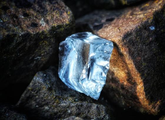 A close-up of a weathered piece of broken glass, roughly cubic in shape, resting among dark, textured rocks. The glass has a translucent, light blue tint with uneven, jagged edges. The surrounding rocks appear rugged and are partially covered in patches of dirt or lichen. The lighting highlights the glass’s sharp edges and internal patterns, creating a stark contrast against the natural, earthy tones of the rocks.
