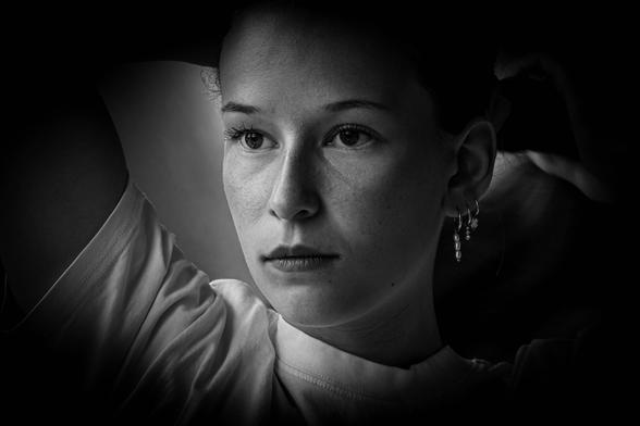 Black and white portrait (face only) of a young woman in low-key. She's looking slightly to the left (actually, to the right!) in the photo, holding her hair up with her hand above her head. Only the left half of her face is illuminated, as is part of the arm that is holding up her hair. A little light also falls on her earrings and right ear. So, the left part of the face is illuminated, and the right part is much darker except for the ear.