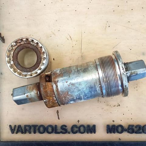 Photo format carré présentant un boîtier de pédalier de vélo, à axe carré, posé sur une surface claire : Un cyclindre métallique, très corrodé, se terminant à chaque extrémité par un emmanchement carré. La partie droite du cylindre présente un filetage pour le visser. Du côté gauche, la rouille est venue à bout du roulement scellé qui a cassé et laisse les billes apparentes.
Au-dessus du cylindre, un objet métallique, rouillé aussi, de forme ronde.