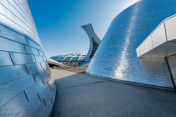 Montreal's Olympic stadium seen in the distance between the twin domes of the Alcan planetarium under a clear blue sky. The shiny metal tiles reflect the blue sky giving the entire image a cyan tint giving the impression everything is bathed in sunlight.