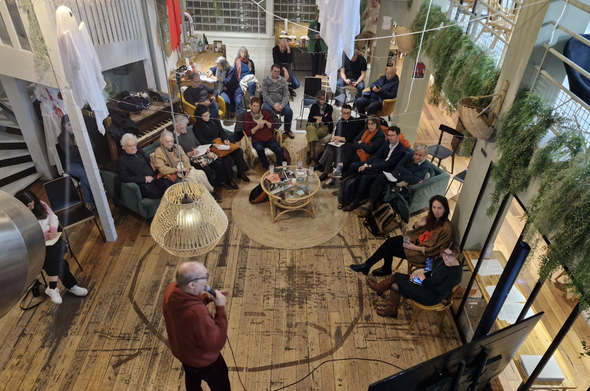 Photo d'ambiance prise de haut, montrant (en bas de la photo) David Cormand tenant un micro et discourant à cÎté d'un écran, devant un parterre d'une petite trentaine de personnes assises sur des canapés et des chaises, autour d'une table bases en osier.