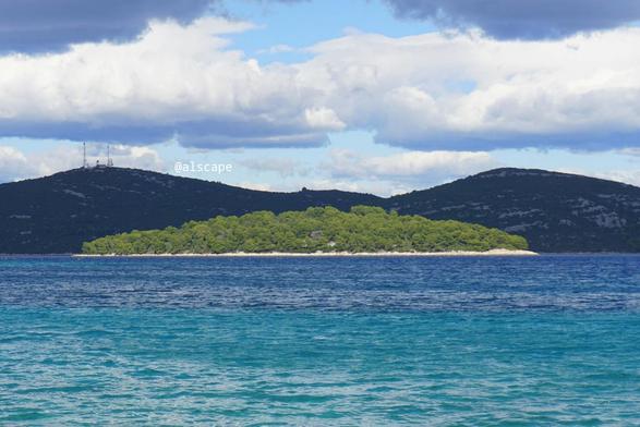 Fotografia przedstawiaj膮ca jedn膮 z wysp w pobli偶u wyspy Murter. Zosta艂a zrobiona w艂a艣nie z tej wyspy. Za t膮 wysp膮 mo偶e by膰 archipelag Kornat lub pasmo g贸rskie kontynentu Dalmacji, nie wiadomo. Wyspa znajduje si臋 w centrum zdj臋cia. Kolor morza pionowo pod wysp膮 jest w wi臋kszo艣ci turkusowy. Morze jest spokojne chocia偶 drobne fale przez lokalne podmuchy s膮 widoczne. Wyspa w wi臋kszo艣ci jest poro艣ni臋ta zielon膮 ro艣linno艣ci膮. U jej podn贸偶a widoczne jest niewysokie pasmo skalistego brzegu koloru kremowego. Niebo nad wysp膮 jest w po艂owie pochmurne, ale jest bardzo jasne.