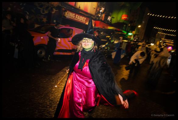 Young woman in elaborate witch costume with black pointed hat, green face paint, hot pink satin dress and black cape poses during Dragon of Shandon parade on North Main Street Cork at night with pink and orange striped van and colourful murals visible behind.