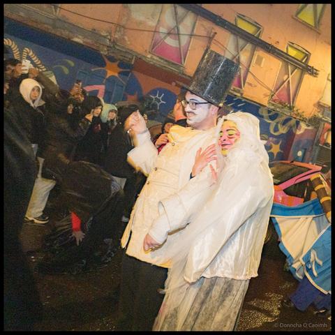 Parade participant in cream-coloured elaborate Victorian-style coat with tall black top hat and theatrical face paint holds yellow fabric puppet during Dragon of Shandon parade on North Main Street Cork with colourful mural backdrop.
