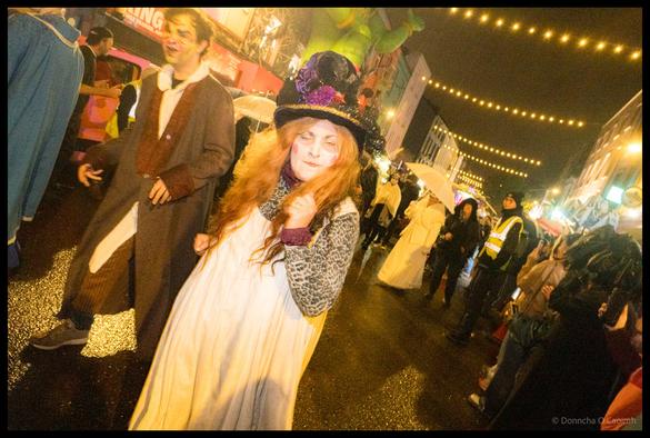 Young person with long red hair in Victorian-style white dress, leopard print shawl and ornate black top hat with purple flowers poses during Dragon of Shandon parade on North Main Street Cork under string lights at night