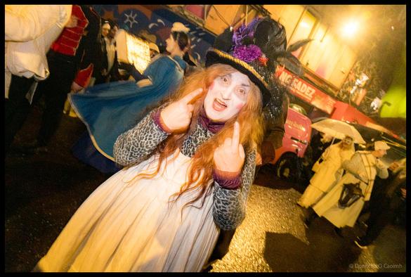 Participant with long red hair in Victorian dress, black decorated top hat and theatrical face paint makes peace signs with both hands during Dragon of Shandon parade on North Main Street Cork with King Fadez shop visible in background