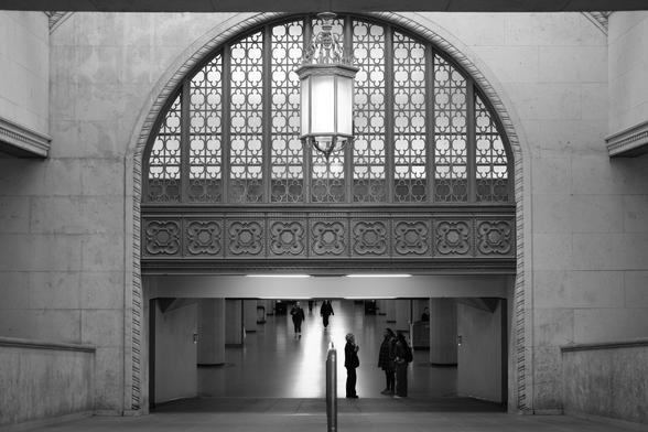 Looking down the ramp to the curved arch leading to the VIA Rail concourse.  Above is an intricately designed fan light surrounded by carved patterns in the stone. In the opening are framed various customers and employees, small in the distance. Down the centre of the concourse are two bright lines of reflected light, leading into the distance.