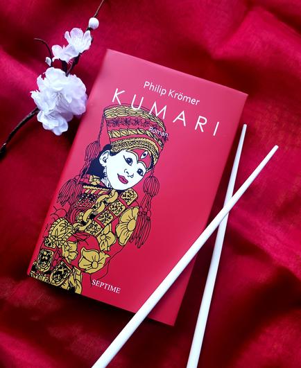 Foto des Buches, auf dessen knallrotem Cover die Zeichnung einer Kumari zu sehen ist – eines stark geschminkten Mädchens mit asiatischen Gesichtszügen und in prächtigen, ornamentierten Gewändern mitsamt einer passenden Kopfbedeckung. Das Buch liegt auf knallrotem, glänzendem Stoff, links oben daneben eine weiße Kunstblume (vielleicht sollen es Apfel- oder Kirschblüten sein), rechts daneben ein Paar weiße Essstäbchen.