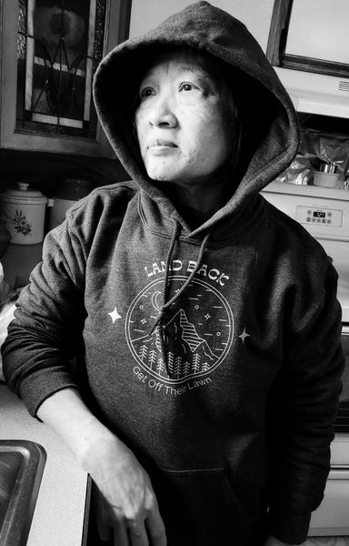 Black and white photo of an Asian person in a hooded sweatshirt standing in a kitchen, looking to the side, leaning on a counter. The sweatshirt displays a circular mountain logo with text “Land Back” and “Get off their lawn.” The kitchen includes a window with a decorative pattern, a stove, and various jars and containers.