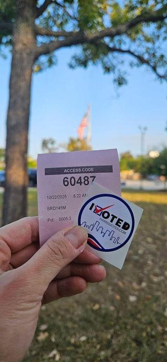 A left hand holding an "I Voted" sticker for Harris County over a receipt with Access Code 60487 dated 10/22/25 at 8:22 AM in the foreground. In the background is grass, a tree, and a blurry American flag flying on a flagpole.