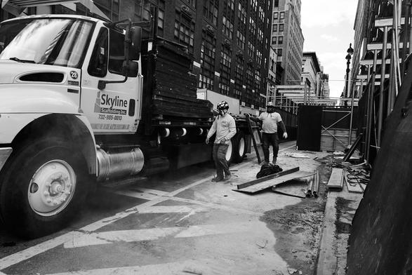 Two construction workers in safety gear walking near a construction truck on a city street surrounded by tall buildings and scaffolding.