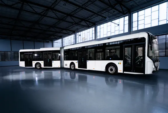 A modern, articulated bus with a sleek design occupies a spacious, brightly lit warehouse. The bus is predominantly white with subtle accents and features large windows, allowing light to stream in. The polished floor reflects the bus, emphasising its length and futuristic appearance.