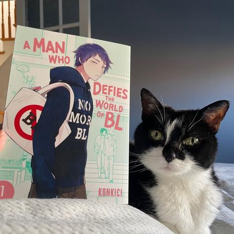 Cute black and white cat shown next to a BL manga volume.